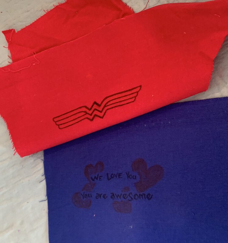 Image shows a blue fabric with a selvedge across the top laid flat on an ironing mat. On it is written "We love you. You are awesome". Above it is placed red fabric with the outline of Wonder Woman's logo drawn on in back. The fabric is folded over as I try to figure out how to line it up to cover the selvedge and not the drawings. 