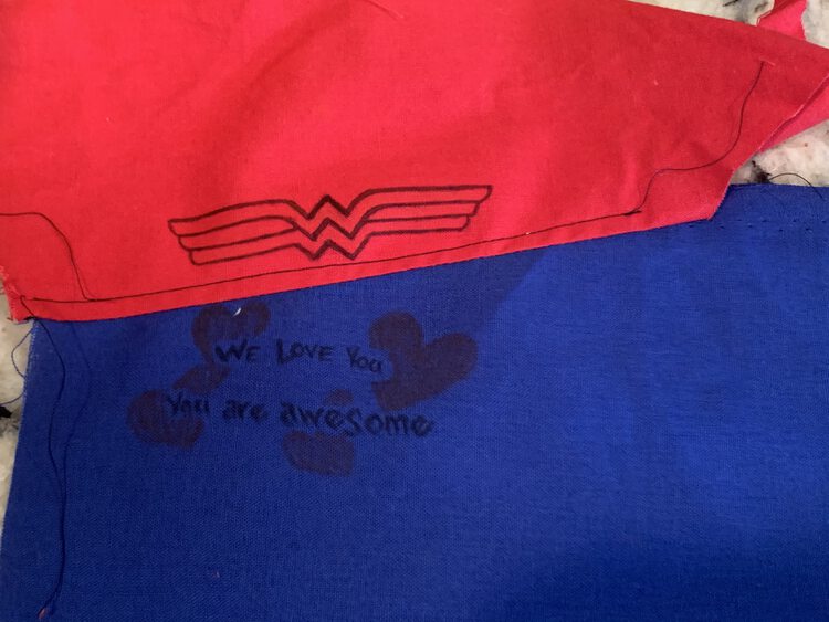 Image shows a red fabric and blue sewn together with a black line of top-stitching along the red fabric to hold the seam down. There's an outline of Wonder Woman's logo on the red fabric and a message on the blue. 