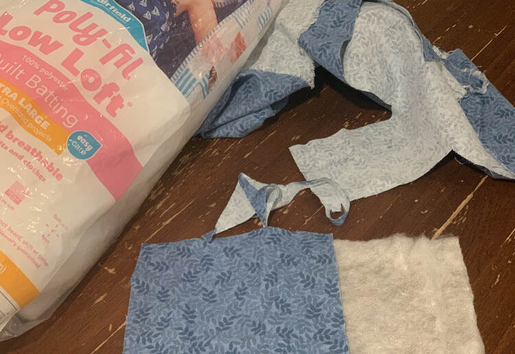 Image shows a a square of quilt packing next to a roughly cut square of blue leafy fabric. Behind it sits a package of poly-fil low loft quilt batting along with the scraps from the snuggle flannel.