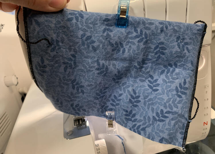 Image shows a rectangle of fabric being held up in front of a serger. There's a clip at the center top and bottom of the rectangle. The sides have a rolled hem, the top is folded over, and the bottom shos a raw edge.