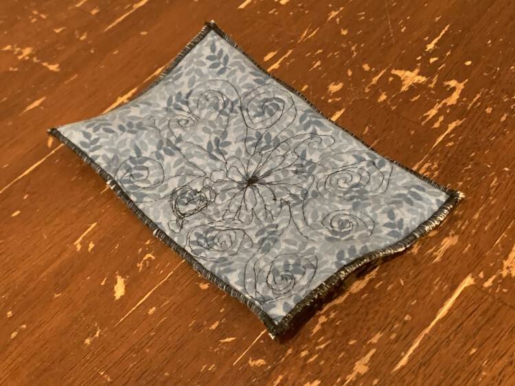 The coaster sits on the kitchen table. You can see a black threaded flower off center on it with black spirals radiating out from it. The edges are finished with a black rolled hem.