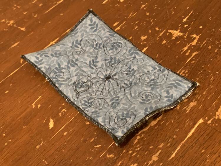 The coaster sits on the kitchen table. You can see a black threaded flower off center on it with black spirals radiating out from it. The edges are finished with a black rolled hem. 