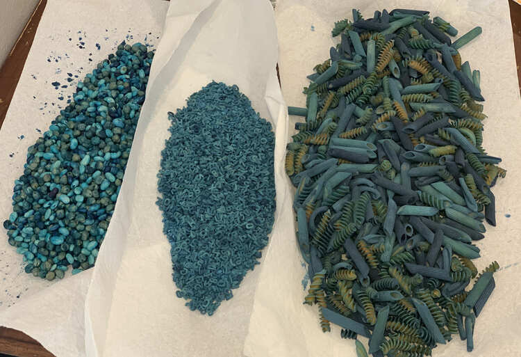 Image shows three piles of blue sensory material laid out on overlapping paper towel. From the left to right it's a mix of large pasta, rice with alphabet pasta, and two types of beans.