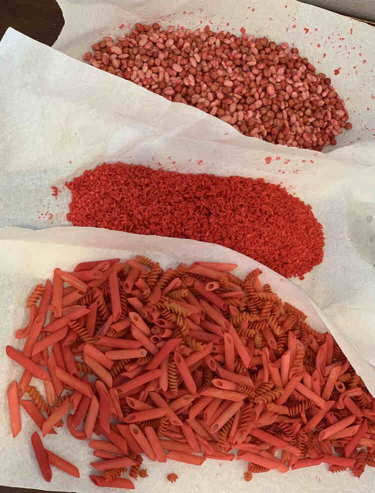 Image shows three piles of red sensory material laid out on overlapping paper towel. From the left to right it's a mix of large pasta, rice with alphabet pasta, and two types of beans. 