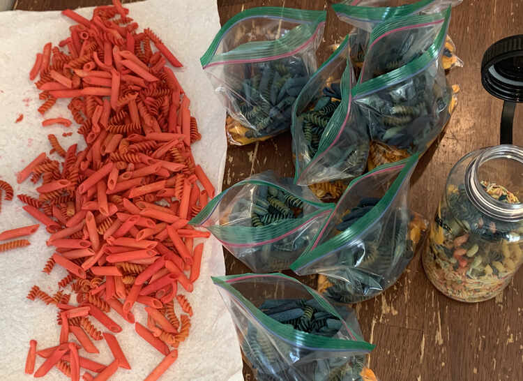 Image shows seven bags of blue and yellow (hidden) pasta laid out in the center of the photo. To the left lays the red pasta on a sheet of white paper towel. To the right lays an opened plastic bottle of faded pasta. 