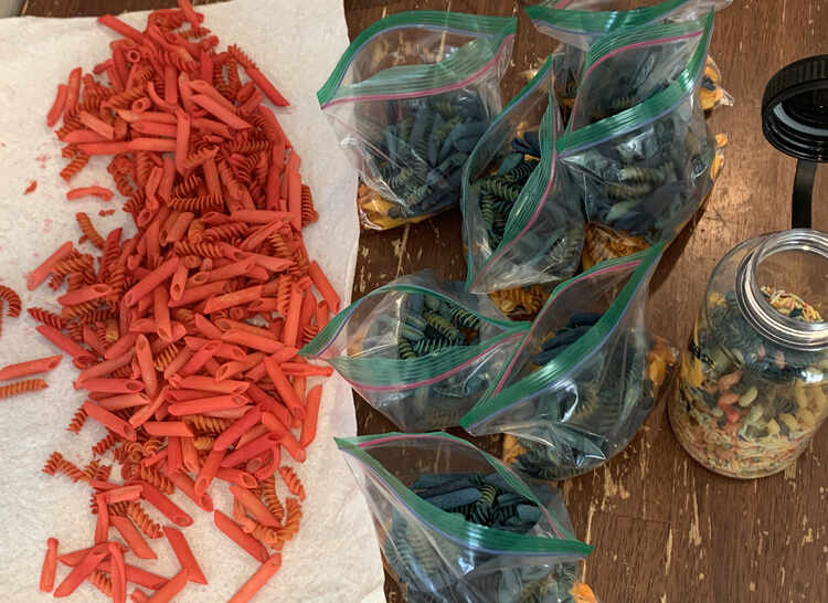 Image shows seven bags of blue and yellow (hidden) pasta laid out in the center of the photo. To the left lays the red pasta on a sheet of white paper towel. To the right lays an opened plastic bottle of faded pasta.