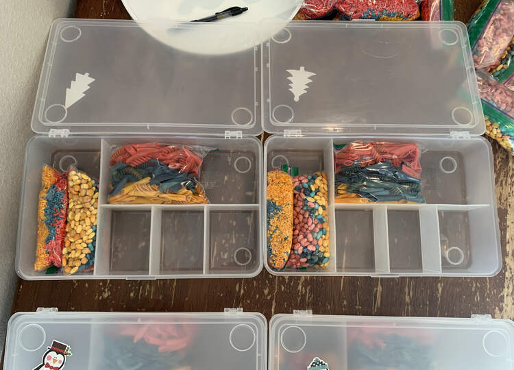 Image is taken from above looking at partially filled sensory bins. The left section has two multicolored bags of dyed alphabet pasta/rice and beans. The top portion has a sandwich-sized bag with dyed pasta. The other sections are empty. At the bottom of the image you see a portion of two closed sensory bins while the excess bags of dyed material are around the top. 
