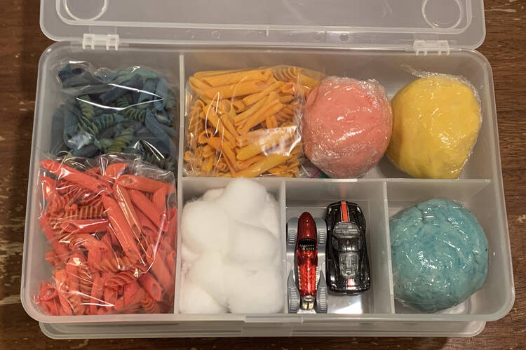 Image shows a sensory kit geared towards a younger kid. In it sits three bags of dyed rotini and penne pasta, three bags of dyed playdough, one section of fluffy white cotton balls, and two toy vehicles. 