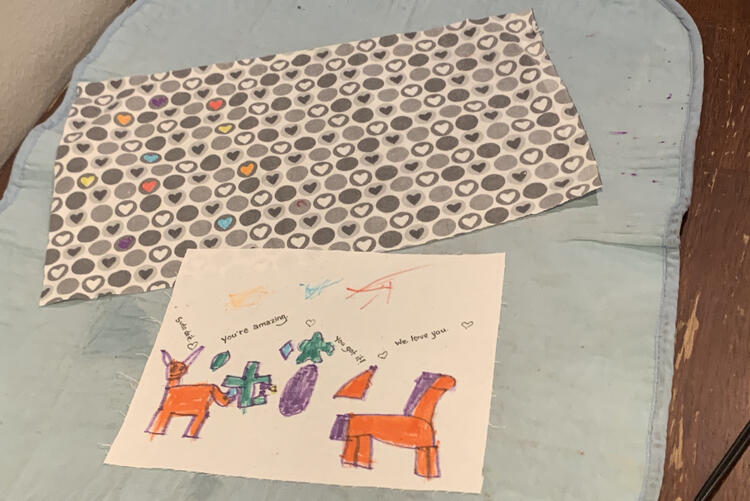 Image shows both fabrics laid out on the ironing mat at the kitchen table. The heart fabric has the odd heart colored in with yellow, red, orange, blue, or purple ink while the white fabric has unicorns, shapes, scribbles, and messages. 