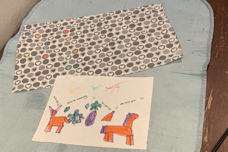 Image shows both fabrics laid out on the ironing mat at the kitchen table. The heart fabric has the odd heart colored in with yellow, red, orange, blue, or purple ink while the white fabric has unicorns, shapes, scribbles, and messages.
