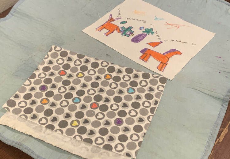 Image shows the grey and colored in heart fabric folded in half so the bottom edge sticks out more than the top showing the underside of the selvage. Behind it sits the decorated white fabric. Both sit on a blue ironing mat on the kitchen table.
