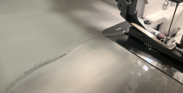 Image shows a close up of the white thread tail behind the serger's foot. The slightly closer end is wider and blurred while the thread closer to the foot has switched to a skinnier stitching. 