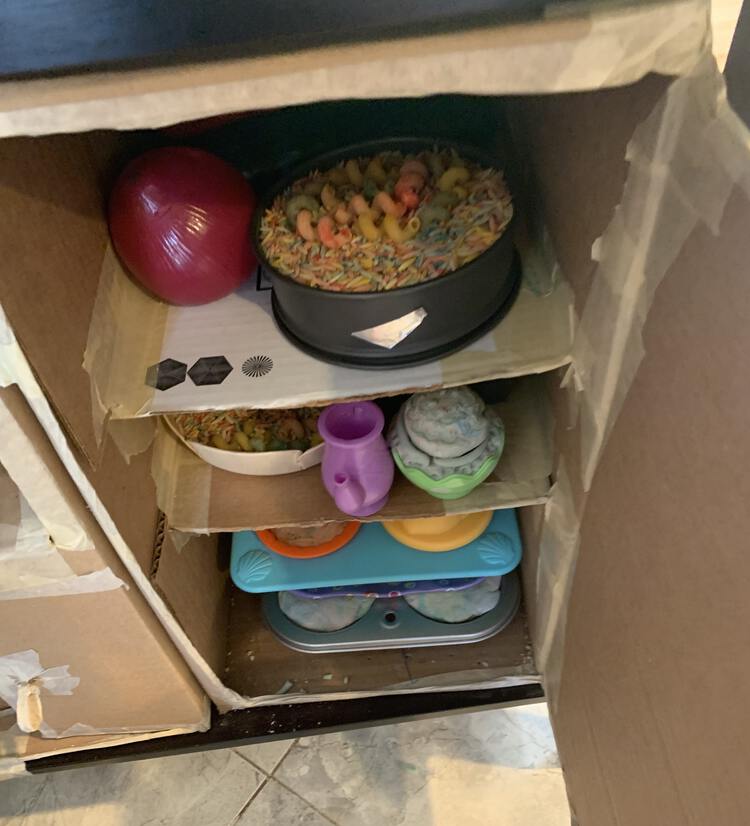 Image shows a closeup of the opened shelf lined cardboard the represents a fridge in our kitchen set. The top shelf has a cake pan filled with vinegar-dyed pasta topped with corkscrew pasta sitting beside a plastic onion. The second shelf sports a half egg shell filled with blue playdough cake, a plastic teapot, and a wooden brie tray filled with pasta. The last shelf shows staked muffin trays filled with playdough cupcakes. 