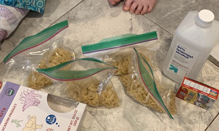 Image shows four plastic sandwich bags sitting on the tiled floor equally filled a quarter way with dinosaur pasta. Around it sits the emptied box of pasta, a bottle of 91% isopropyl alcohol, a package of four neon water-based food dyes, and part of my kids as they sit beside it all. 