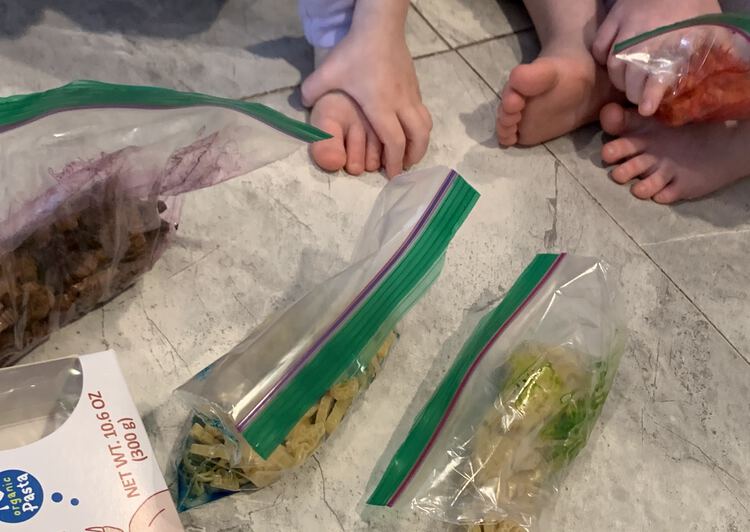 Image shows a mixed sealed bag of purple pasta on the left, two bags of unmixed dyed pasta in the center, and a red bag of pasta in the corner being mixed by a kid. 