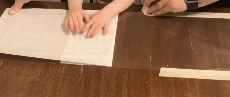 Image shows Ada helping to hold the backing from the con-tact paper down while I attach masking tape to the sides of the stick clear paper.