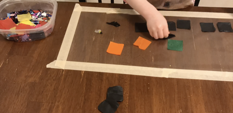 Image shows the taped down clear contact paper with several black squares and some other colored squares of tissue paper on it. There's masking tape around the outer edge holding it to the table and a container of more tissue paper squares and confetti next to it. 