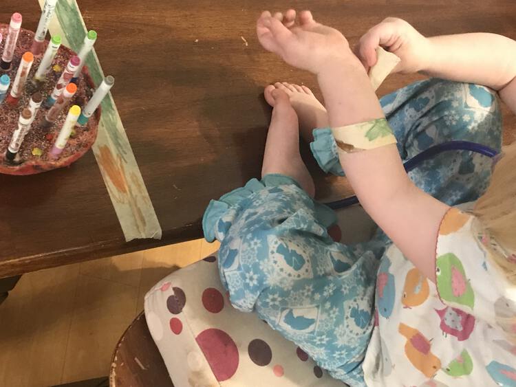 Image shows Ada sitting on her chair in front of the bench wrapping a decorated masking tape bandage around her arm. In the background are the other stripes of tape and the felt pens. 