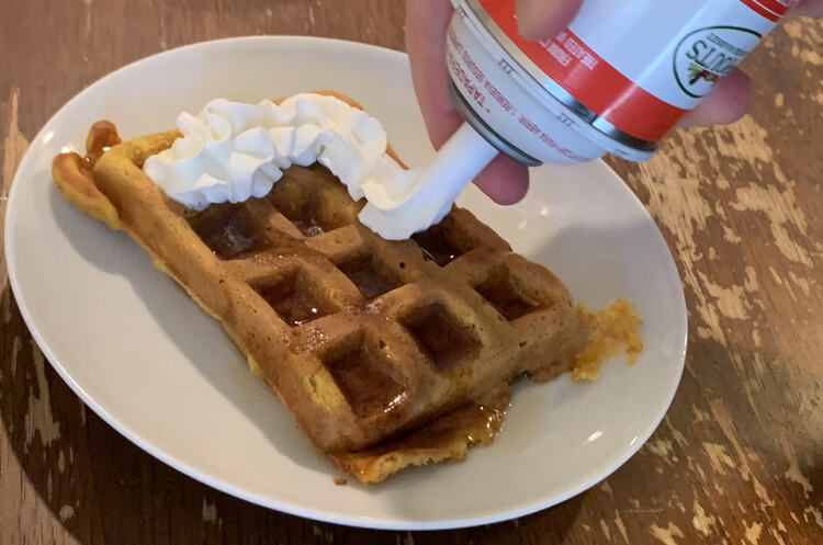 Image shows a single waffle on a white saucer with wet syrup on parts and a stream of white whip cream coming from a canister. 