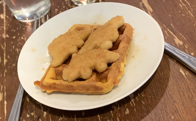 Image shows a waffle with a t-rex, stegosaurus, and triceratops shaped chicken nuggets over top. 