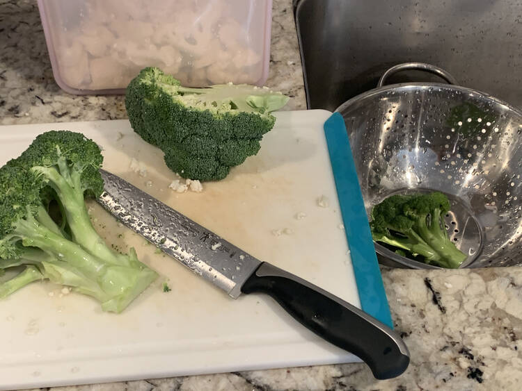 Image shows a plastic cutting board with a knife and two half stalks of broccoli. In the sink beside it there's a metal strainer with a small broccoli stalk. Behind the cutting board is a pink transparent Stasher bag with cut up cauliflower inside it. 