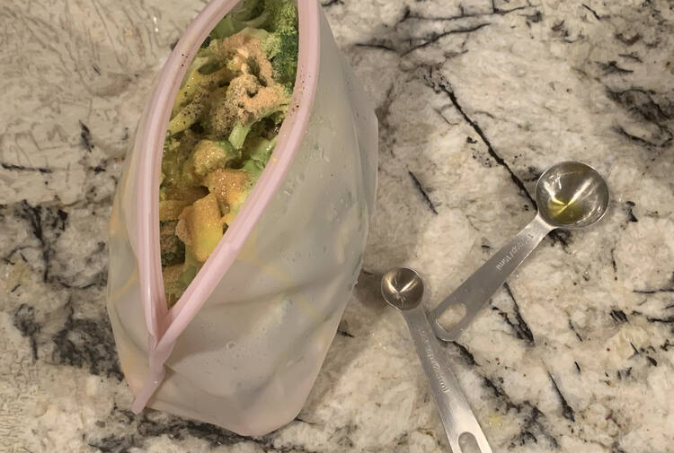 Image is taken from above looking at the opened stasher bag showing broccoli with the other ingredients over top of it. Beside it sits two measuring spoons. 