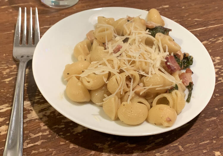 Image shows Pipe Rigate mixed with bacon, swiss chard, beans, and topped with grated cheese. It's plated on a white saucer with a fork next to it. 