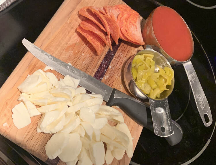 Image shows a wooden cutting board with diced mozzerella, some a knife, frozen pepperoni, a third cup of pepperoncinis, and a half cup of tomato sauce. 