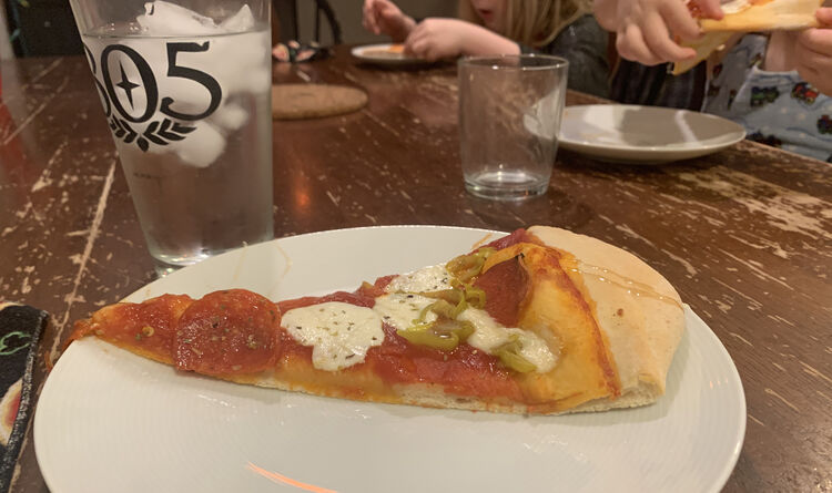 Image shows a single slice of pizza on my white plate. In the distance you can see the kids eating theirs off their plate while Matt is hidden in the back behind my glass. 
