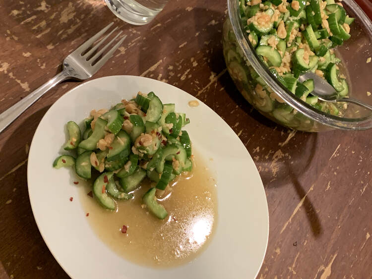 Image shows a saucer with a pile of cut cucumber sitting in a pool of brown liquid. In the background sits a large clear serving bowl with more. 