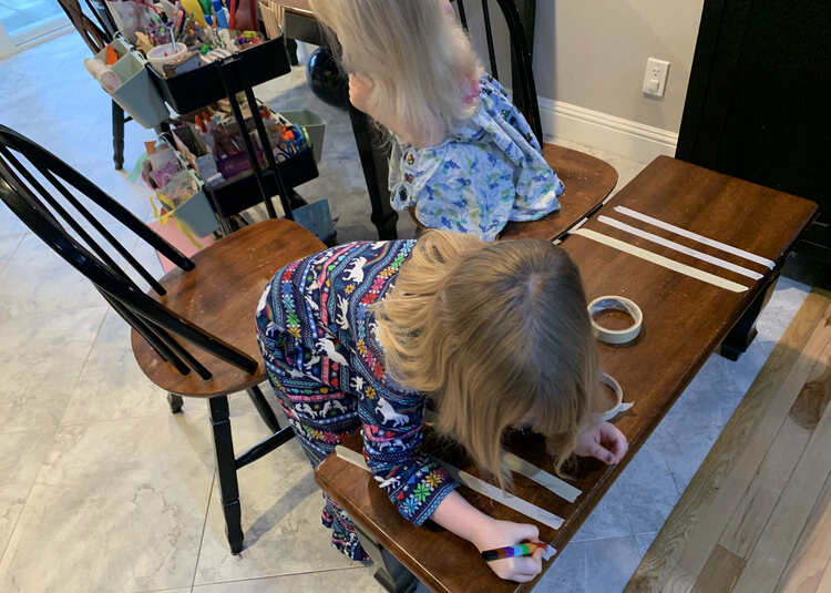 Image shows two chairs pulled up to the bench. Ada leans over her strips of tape while decorating them with a stacked crayon while Zoey is crouched in her chair and turned away from her strips as she peruses the art trolley for supplies.