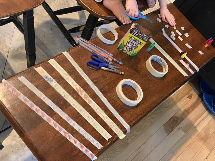 Image shows the bench festooned with many masking tape bandages. Closer to the camera are Ada's patterned strips while, in the background, you can see Zoey's hands and feet while she leans over to cut the tape smaller. 