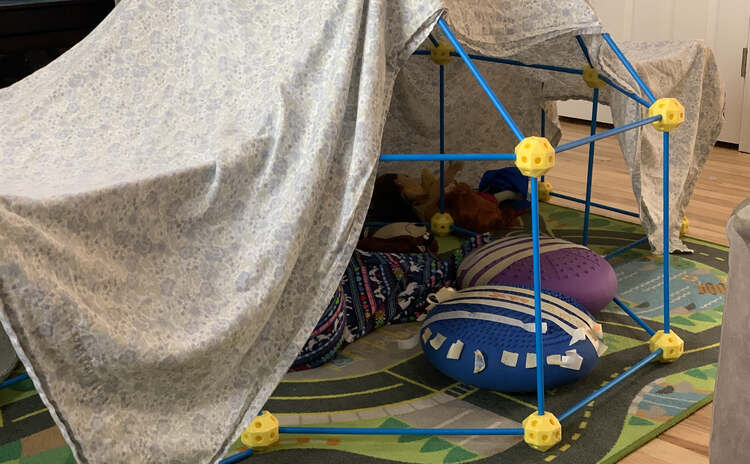 Image shows a ball and stick fort mostly covered in a top sheet. Inside are two wobble cushions covered in decorated masking tape bandages. You can see parts of one kid's nightgown as she doctors her toys. 
