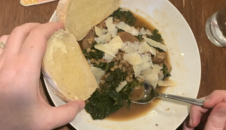 Image shows a white bowl half filled with the same soup as above. This one is coated with slivers of white cheese and has two pieces of white bread to the side. 