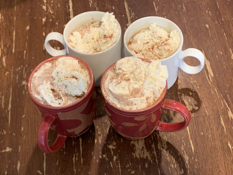 Image shows four cups (two red and two white) sitting on a table. All are filled with hot chocolate, topped with whip cream, and sprinkled with extra toppings. 