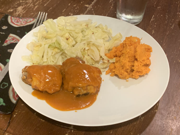 Image shows a white plate with a pizza-themed napkin and fork off to the side. On the plate sits two orange drenched chicken thighs, a mound of orange yams, and a pile of green-ish slivered cabbage.