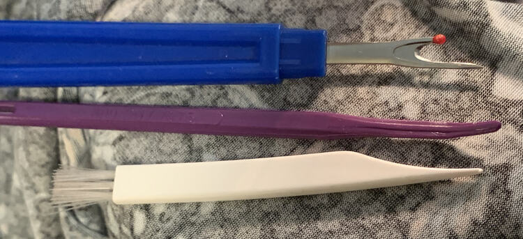 The image shows three tools laid out over the wrong side of some fabric. The top one is my blue handled seam ripper, followed by my That Purple Thang, and then my lint brush that came with both my sewing machine and serger. 