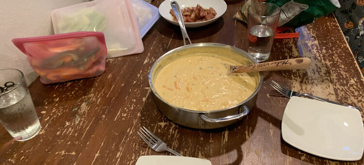 Image shows a metal skillet filled with queso, several stasher bags filled with cleaned and cut up raw vegetables, a bowl of bacon bits, and a bag of tortilla chips. 