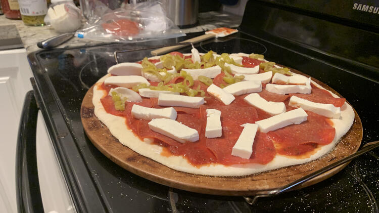 Image shows a freshly decorated pizza about to go into the oven with the ingredients for the second pizza remaining in the background.