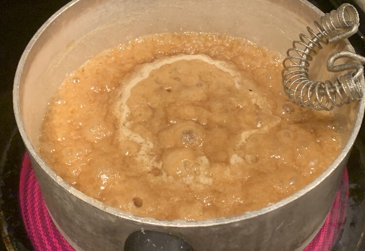 Image shows a bubbling sugar mixture in a grey pan on a red burner. A simple flat bottom whisk sits propped against the pot.