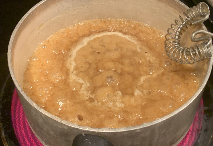 Image shows a bubbling sugar mixture in a grey pan on a red burner. A simple flat bottom whisk sits propped against the pot. 