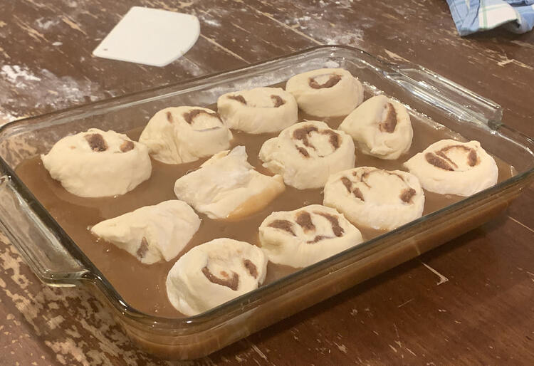 Image shows twelve cinnamon buns sitting in caramel in a glass casserole dish. The pan sits on a hidden cork trivet on a flour splattered table.
