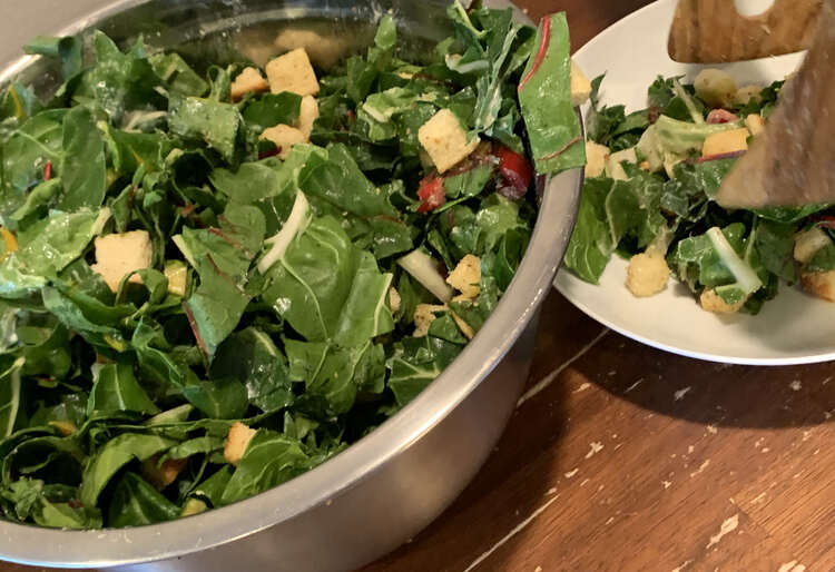 Image shows salad tongs dishing some salad onto a white saucer from a metal bowl. 