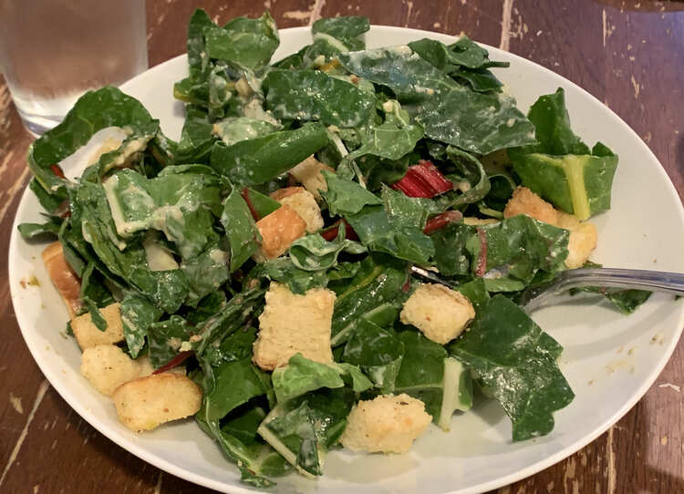 Image shows a wide open bowl filled with Caesar salad, a fork, and a glass of water in the background. 