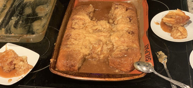 Image shows a cookie sheet with ten cinnamon buns on it as two were dished onto the sauces to the left and right. On the left side you can see the emptied glass casserole dish while at the front there's a fork and spoon for dishing and scooping the buns and sauce. 