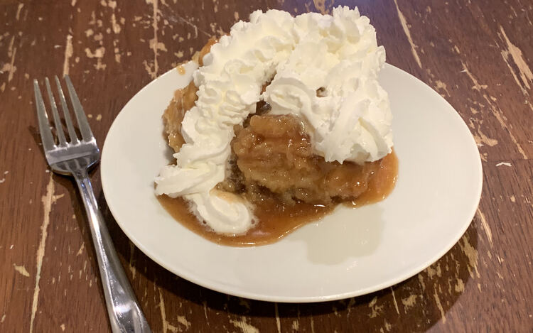 Image shows a piece of sticky bun on a white plate with a pool of caramel. The bun is coated in whip cream and a fork sits beside the saucer about to be picked up. 