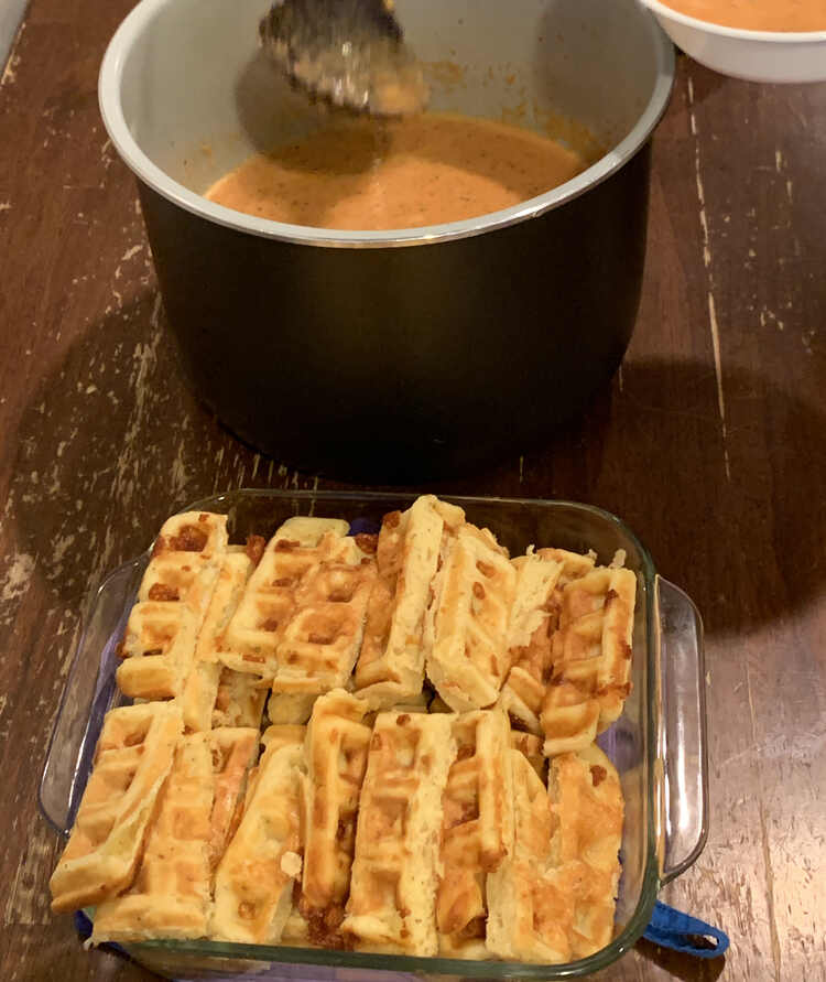 Image shows a pot of tomato soup (in a Ninja Foodi liner) behind a square casserole dish holding the sliced waffle dippers. 