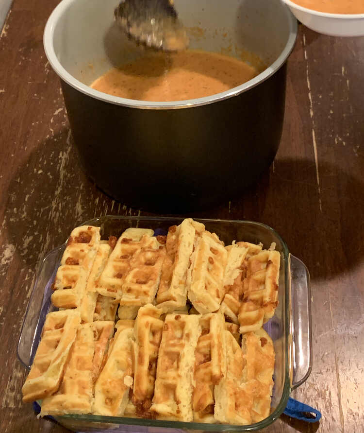 Image shows a pot of tomato soup (in a Ninja Foodi liner) behind a square casserole dish holding the sliced waffle dippers.