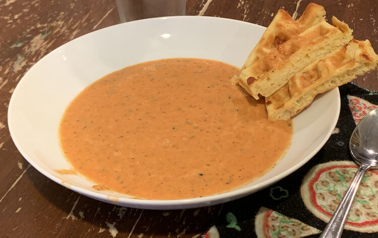 Image shows a white bowl half filled with orange-ish tomato soup and has three slivers of waffles sitting on the edge as if it's dipping it's toes in. Off to the side sits a metal spoon laying on top of a pizza themed napkin.