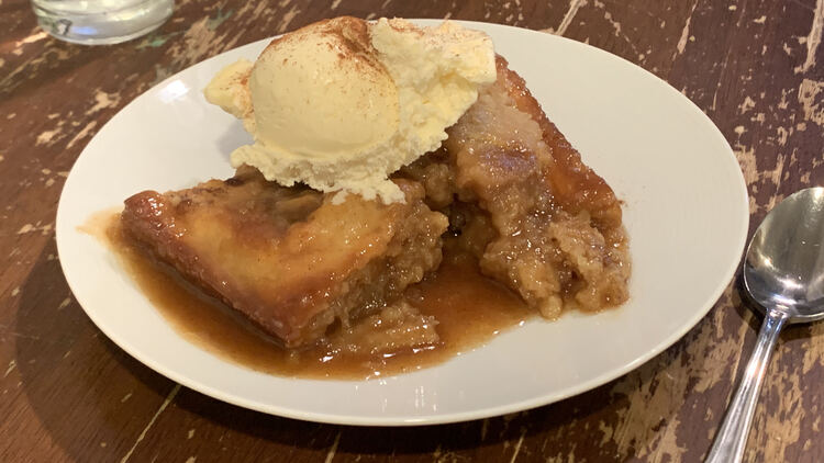 Image shows a decent piece of sticky bun sitting in caramel sauce and topped with a scoop of vanilla ice cream and a sprinkling of cinnamon. This time a spoon sits ready and waiting to be used. 